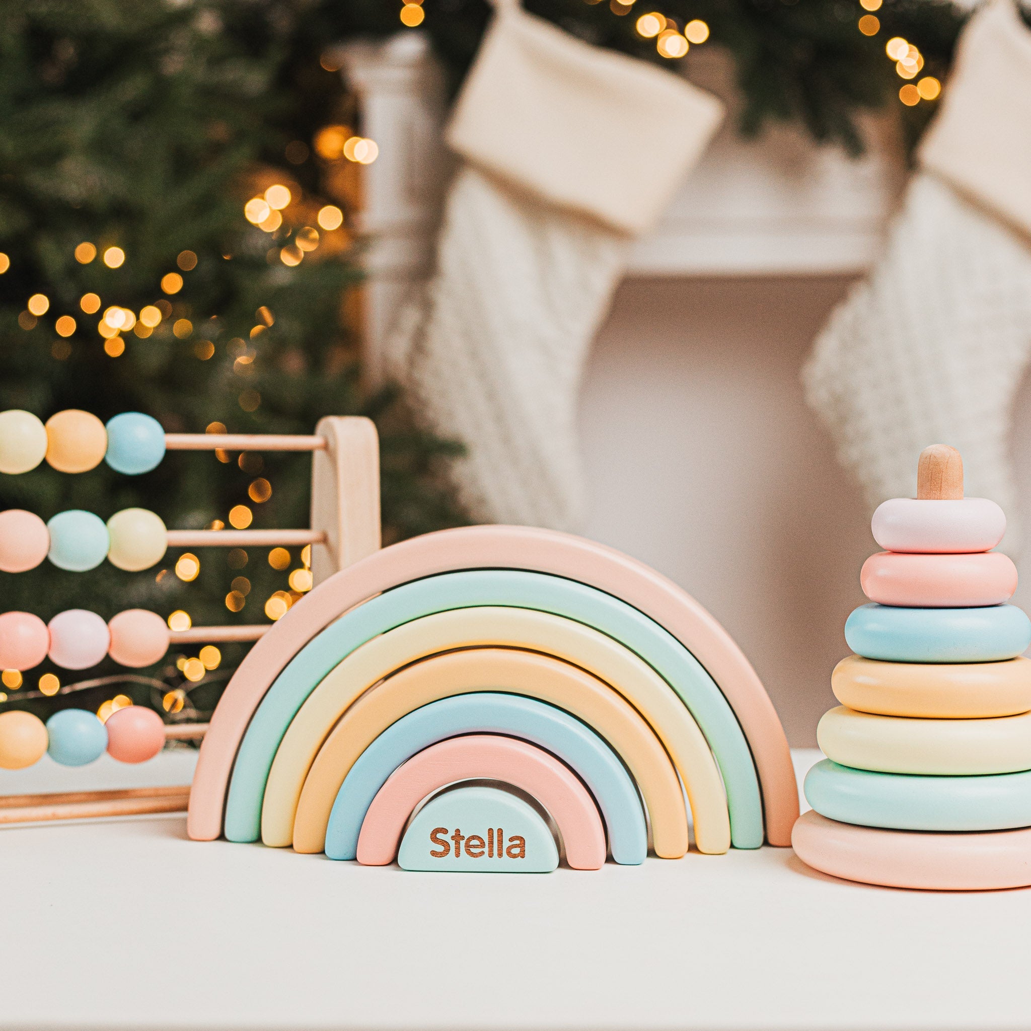 Personalized pastel wooden toys for children, including an engraved rainbow stacker with the name 'Stella,' a colorful abacus, and a soft-toned ring stacker, displayed in a cozy holiday setting.