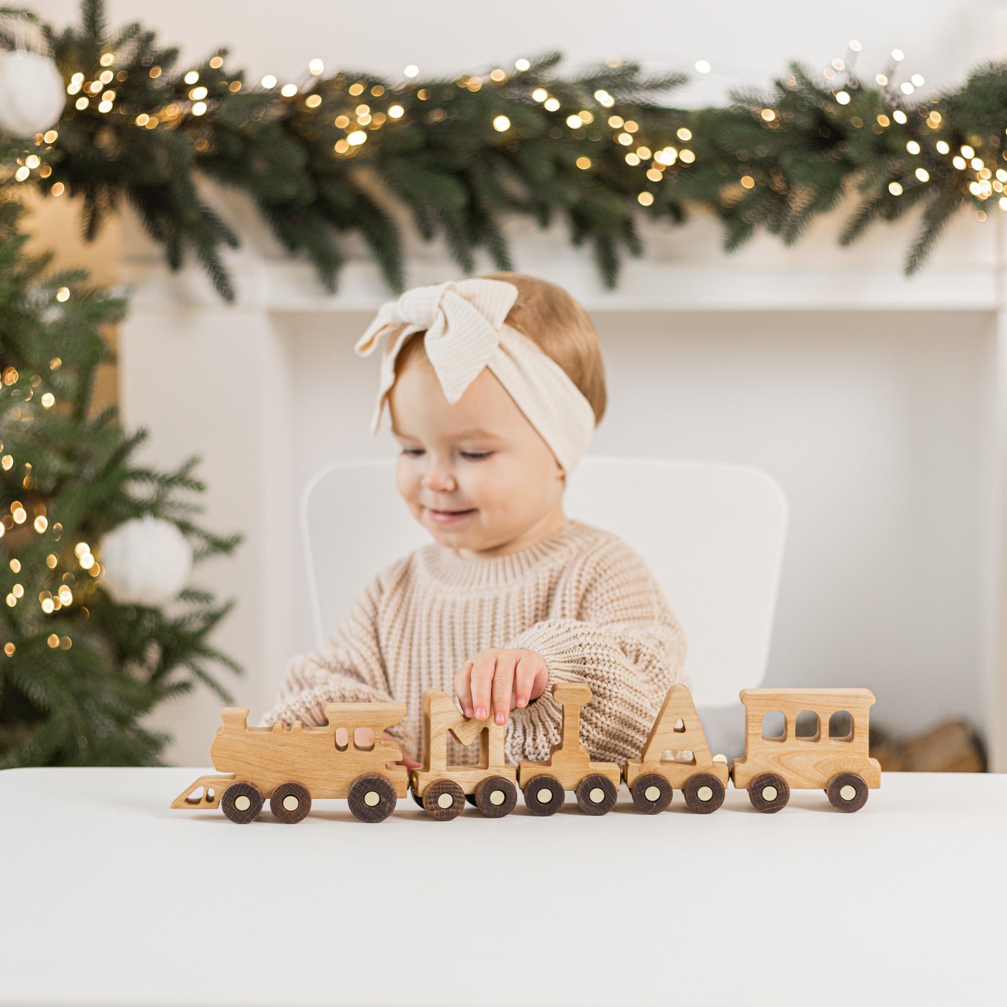 A toddler playing with a personalized wooden name train at a festive table, with a cozy holiday backdrop featuring string lights and evergreen decorations. The train features natural wood tones and customizable letter carriages.