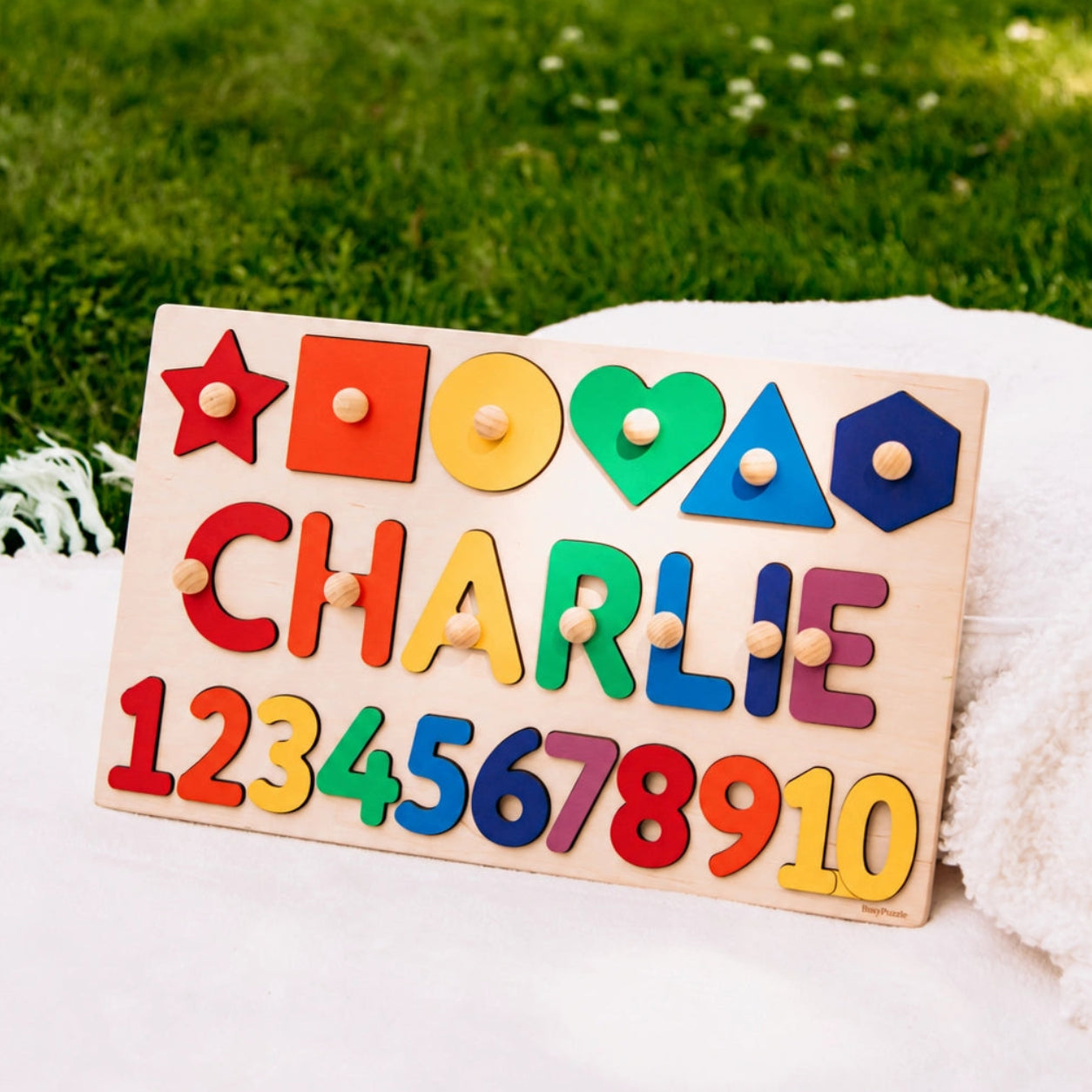 A personalized wooden toy puzzle board with colorful shapes and numbers, customized with the name 'CHARLIE', resting on a white blanket with a green grassy lawn in the background.