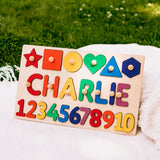 A personalized wooden toy puzzle board with colorful shapes and numbers, customized with the name 'CHARLIE', resting on a white blanket with a green grassy lawn in the background.