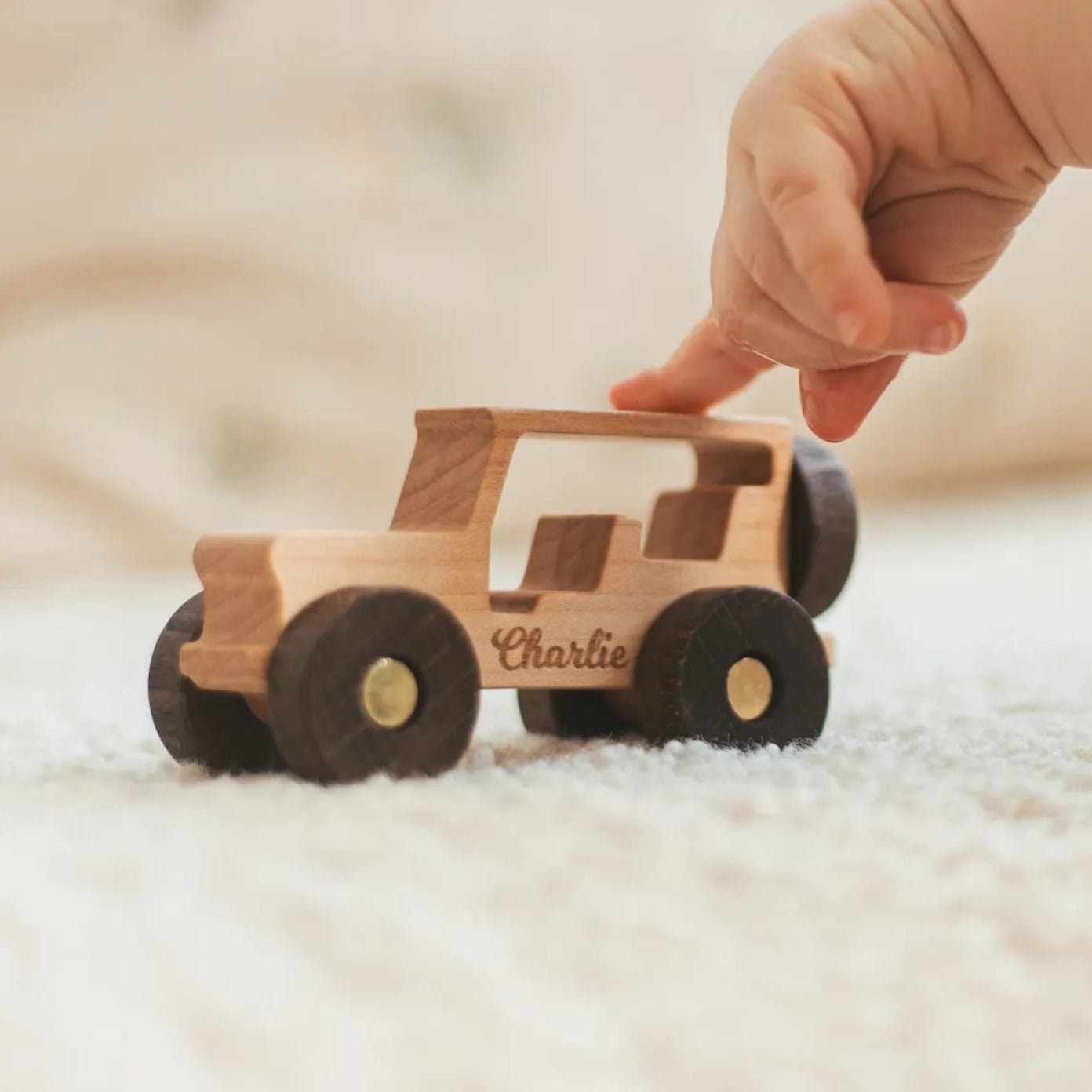 Child's finger reaching toward a natural wood off-roader car engraved with the name Charlie on a soft carpet