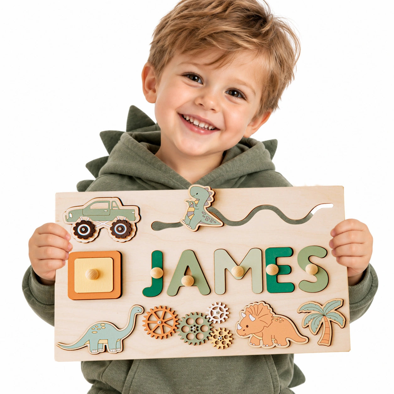 Smiling toddler boy holding a personalized wooden name puzzle with the name JAMES, featuring dinosaurs, truck, gears, and Montessori sensory learning elements.