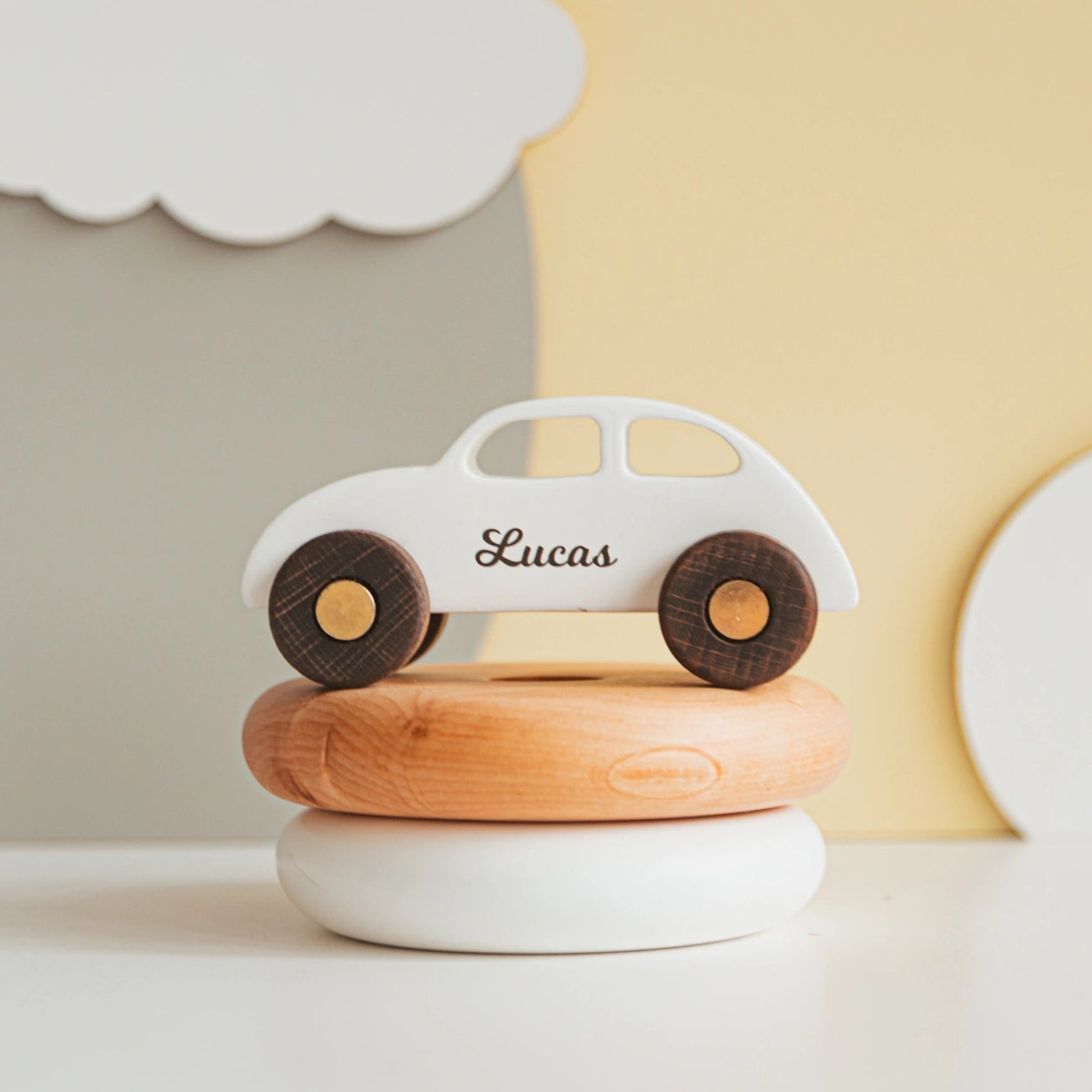 Personalized white wooden coupe car with the name Lucas, resting on a wooden stacking-ring toy in a nursery