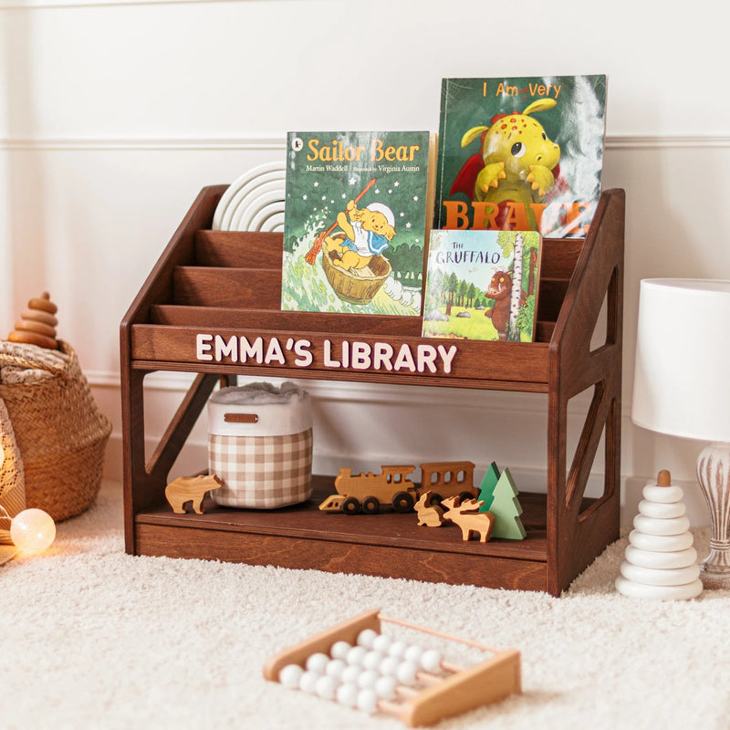 Dark wood personalized Montessori bookshelf with the name Emma's Library, filled with children's picture books in a cozy nursery