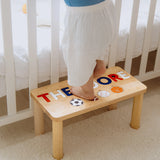 Child standing on a wooden step stool with letters and symbols on a carpeted floor.