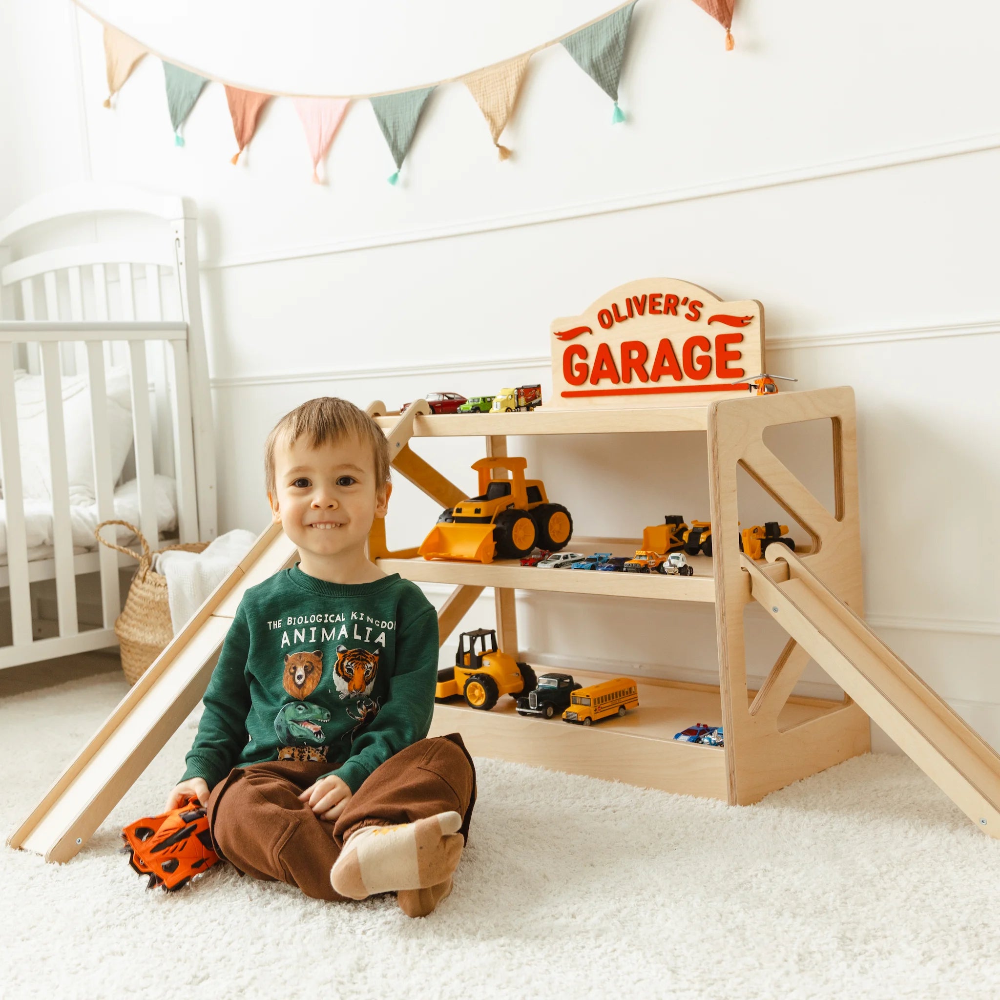 Toddler sitting on the floor playing beside a light wood toy car garage named Oliver's Garage