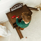 Child playing with toy cars on a wooden play set labeled 'Henry's Garage'.