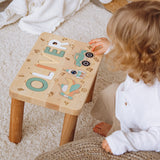 Child playing with a wooden stool featuring cut-out letters and shapes on a carpeted floor.