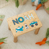 Wooden step stool with name letters and vehicle puzzles on a soft surface.