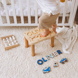 Child standing on a wooden stool with a name on it, surrounded by name blocks and toys in a nursery.