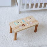 Wooden step stool with personalized name and illustrations on a white carpeted floor.