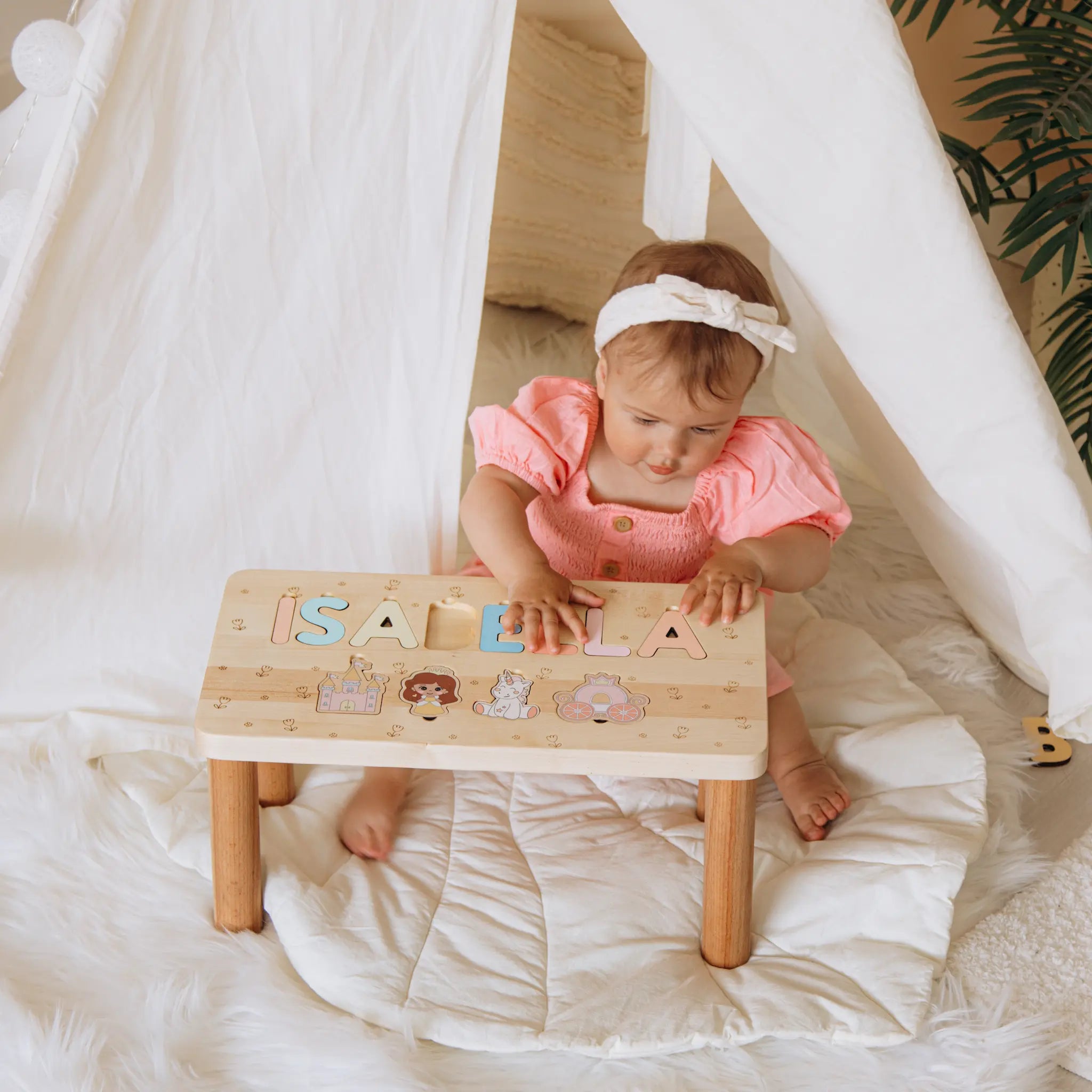 Baby sitting on a wooden stool with letters spelling 'ISABELLA' in a cozy indoor setting.