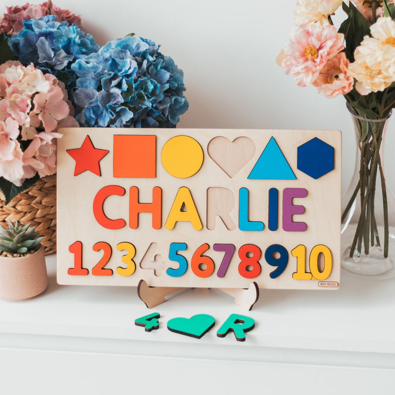 Colorful wooden name puzzle with shapes and numbers on a stand, surrounded by flowers and plants.