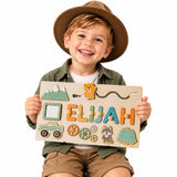 Smiling young boy in a brown hat holding a personalized wooden busy board name puzzle with "ELIJAH", featuring mountain, bear, wolf, forest, truck, and sensory gear elements.