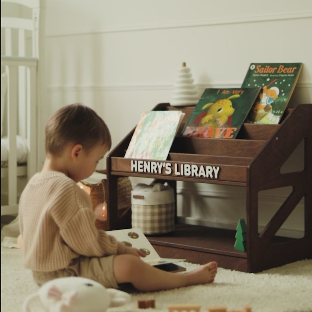 Toddler sitting on the floor reading beside a dark wood personalized bookshelf named Henry's Library